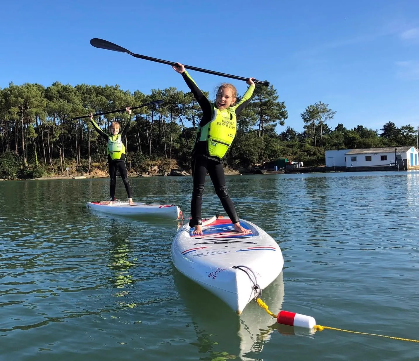 Deux enfants en paddle individuel sur les eaux calmes de la Baie de Quiberon, gilets de sauvetage et pagaies levées