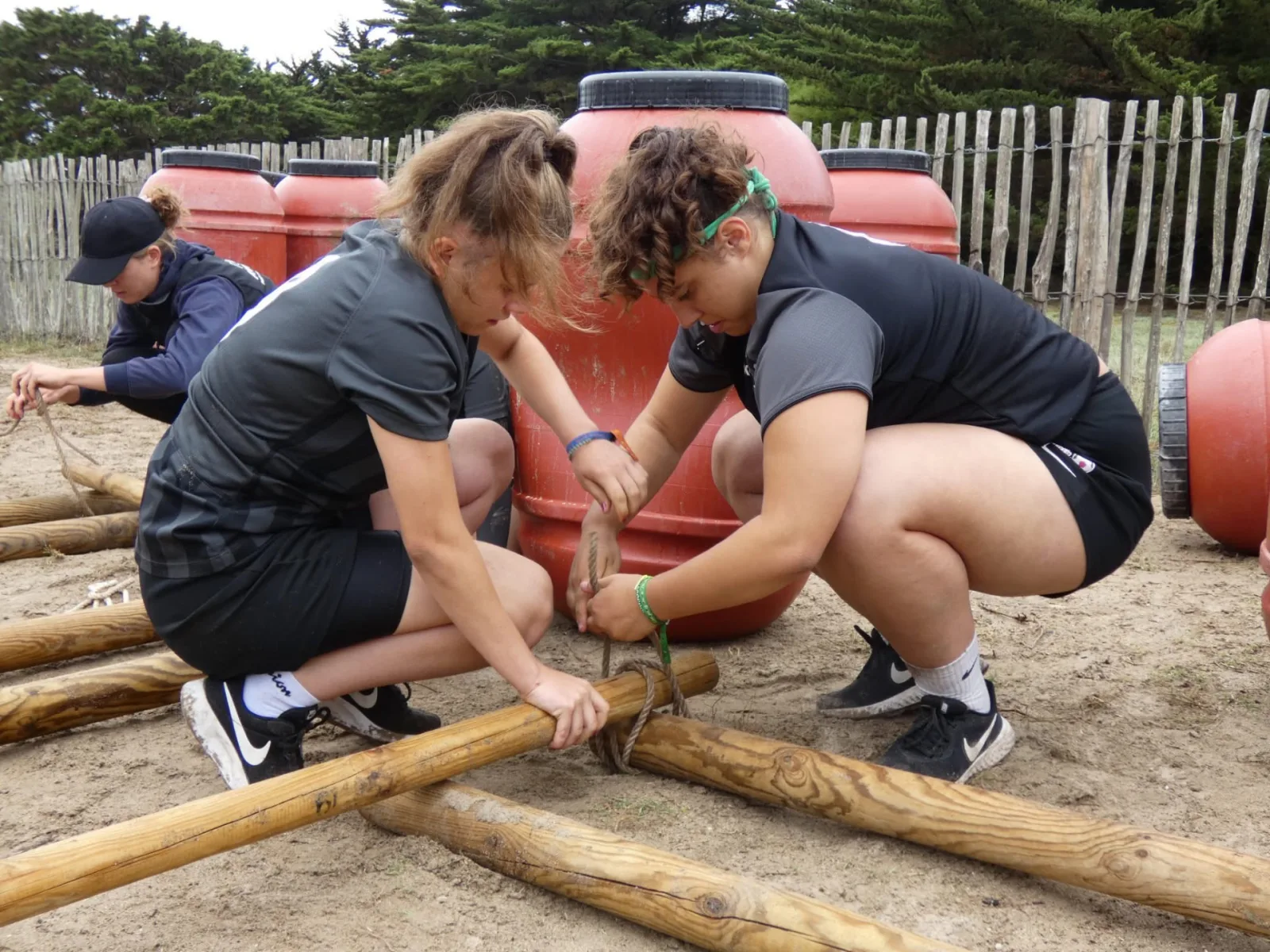 Construction de radeaux en équipe sur la plage de Quiberon : jeunes participantes attachant des rondins de bois et des tonneaux avec des cordes avant la mise à l'eau, presqu'île de Quiberon Bretagne Sud