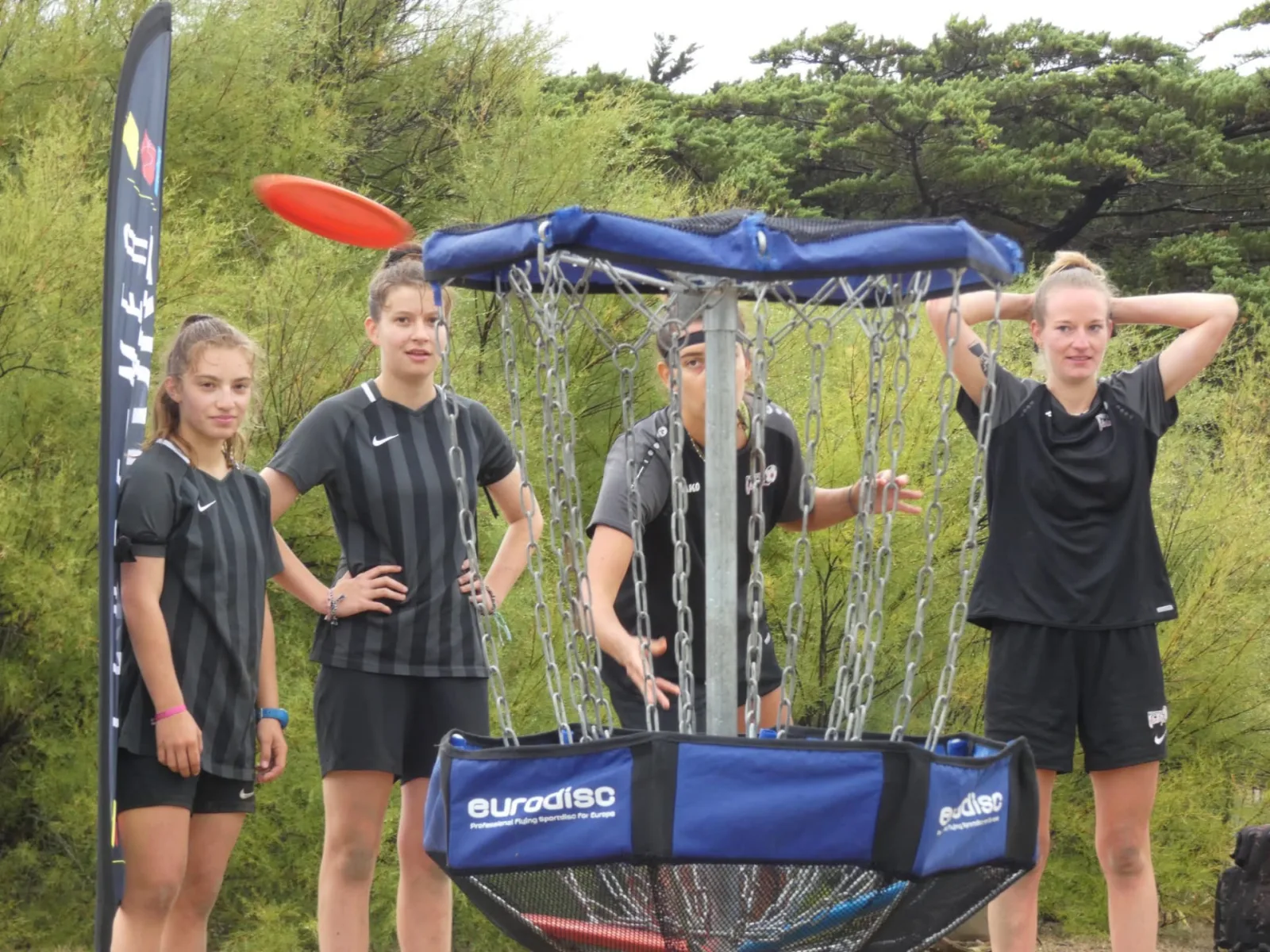 Défi des dunes : jeunes femmes jouant au disc-golf sur la presqu'île de Quiberon, lançant un disque orange vers le panier de réception dans un cadre naturel en Bretagne Sud