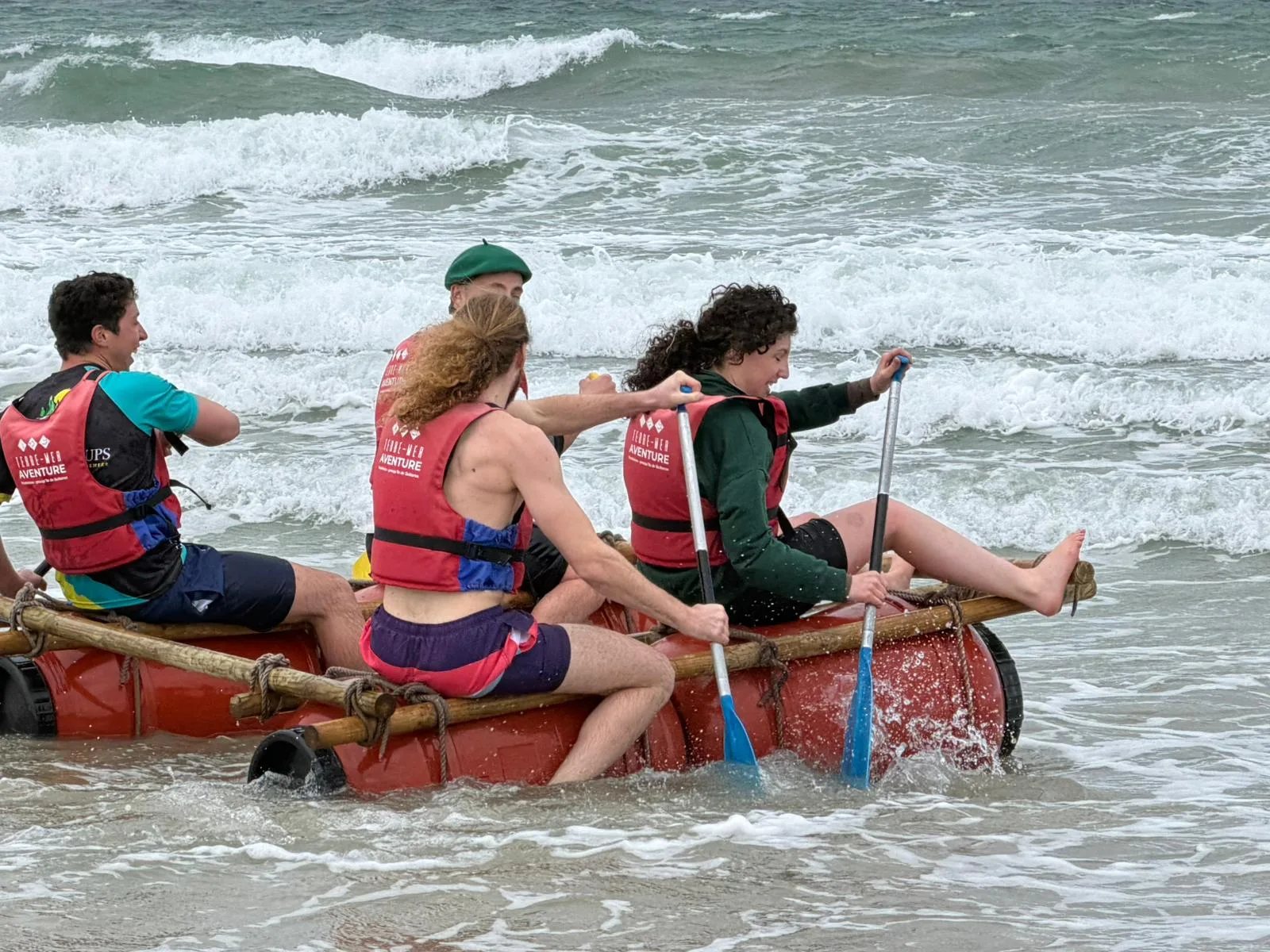 Construction de radeaux en équipe sur la plage de Quiberon : jeunes participantes attachant des rondins de bois et des tonneaux avec des cordes avant la mise à l'eau, presqu'île de Quiberon Bretagne Sud
