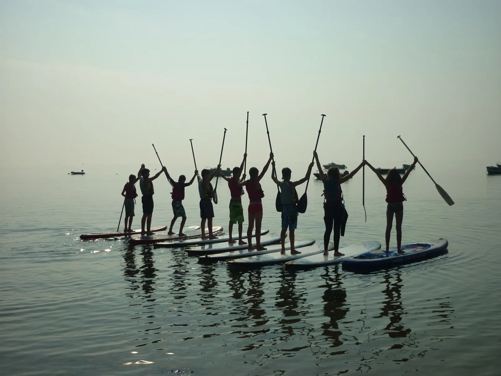Groupe d'enfants en stand-up paddle formant une chaîne humaine et levant leurs pagaies au lever du soleil, Baie de Quiberon Bretagne Sud