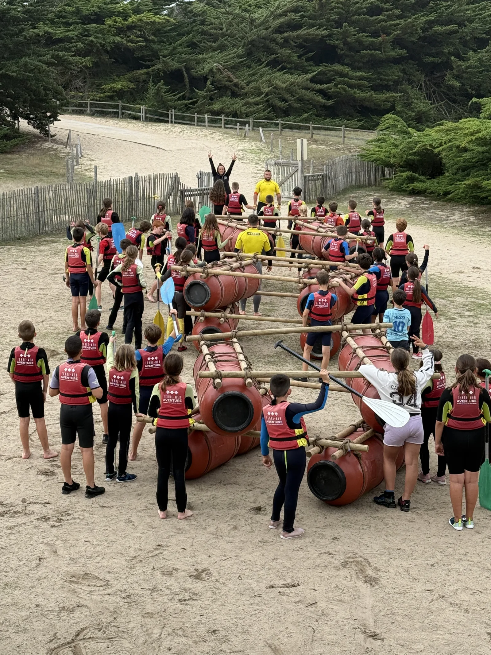 Grand groupe d'enfants en gilets de sauvetage Terre Mer Aventure assemblant radeaux en bidons et perches sur la plage de Quiberon, pagaies à la main